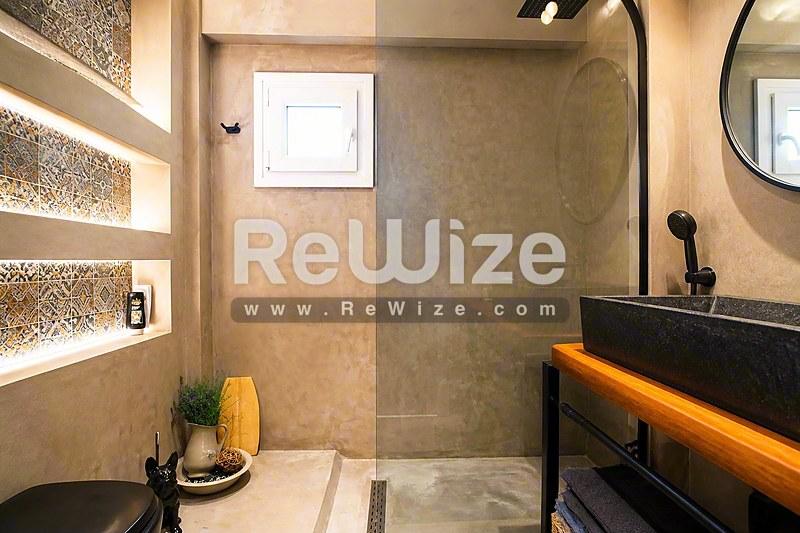 Σύγχρονο μπάνιο διαμερίσματος στη Χαλκιδική με ευρύχωρο ντους, σκούρο νιπτήρα και ζεστό φωτισμό.