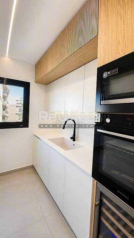 Σύγχρονη κουζίνα σε μεζονέτα στη Χαλκιδική με λευκά ντουλάπια, ξύλινες ανώτερες μονάδες και ενσωματωμένες μαύρες συσκευές.