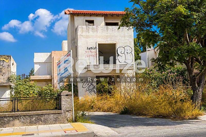 Εγκαταλελειμμένη όψη μονοκατοικίας στη Χαλκιδική με φθαρμένους λευκούς τοίχους και γκράφιτι, περιτριγυρισμένη από άγρια βλάστηση.