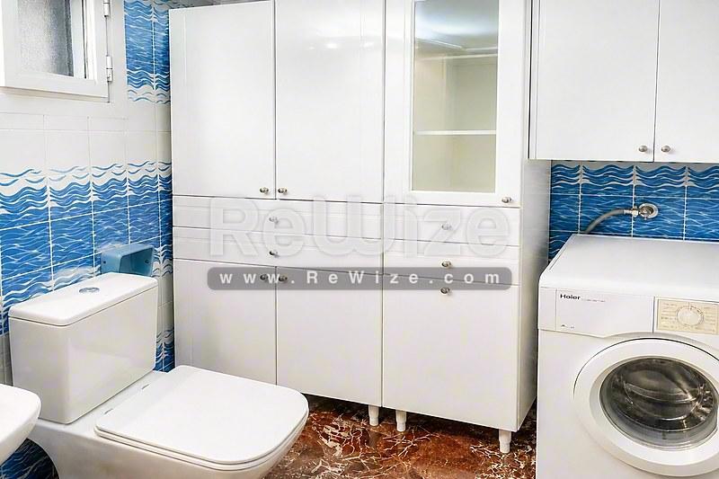 Σύγχρονο μπάνιο στο διαμέρισμα της Χαλκιδικής με λευκά έπιπλα, γαλάζια πλακάκια, τουαλέτα και πλυντήριο.