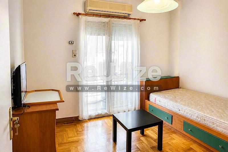 Φωτεινό υπνοδωμάτιο διαμερίσματος στη Χαλκιδική με μονό κρεβάτι, ξύλινα έπιπλα, τηλεόραση και γυάλινες μπαλκονόπορτες.