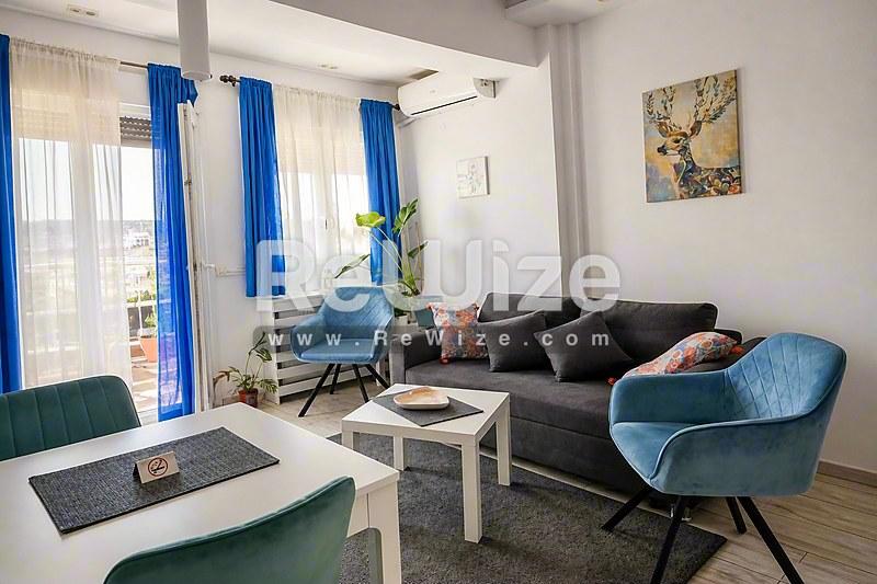 Σύγχρονο σαλόνι διαμερίσματος στη Χαλκιδική με μπλε πολυθρόνες, σκούρο καναπέ και παράθυρα με γαλάζιες κουρτίνες.
