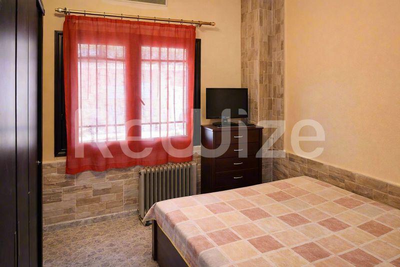 Φωτεινή κρεβατοκάμαρα σε μονοκατοικία στη Χαλκιδική με κρεβάτι, κόκκινες κουρτίνες και ξύλινα έπιπλα.