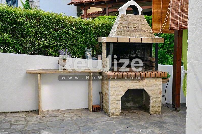 Εξωτερική κουζίνα με τζάκι από τούβλα και ξύλινο πάγκο σε μεζονέτα της Χαλκιδικής, με παραδοσιακό σχέδιο πέτρινου φούρνου.