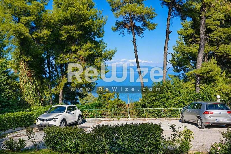 Θέα θάλασσας από μεζονέτα στη Χαλκιδική με γαλάζια νερά, πεύκα και σταθμευμένα αυτοκίνητα.